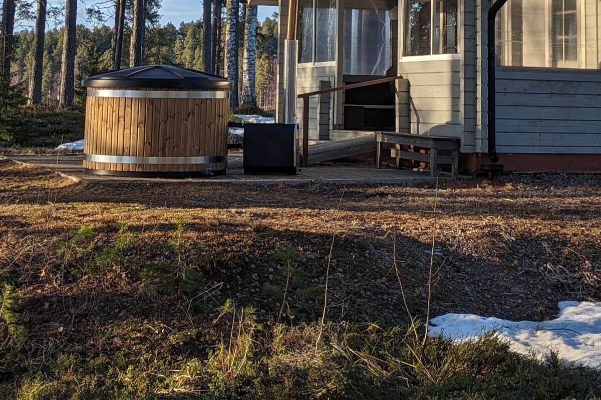 A cabin with a porch, surrounded by tall trees and a hot tub in the foreground.