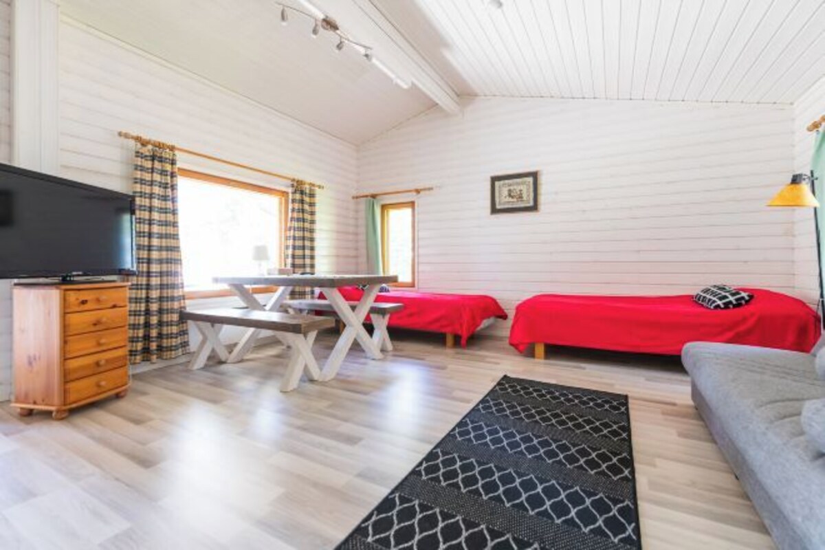 A room with a wooden floor, a TV, a table, two beds with red bedding, a sofa, and a lamp.