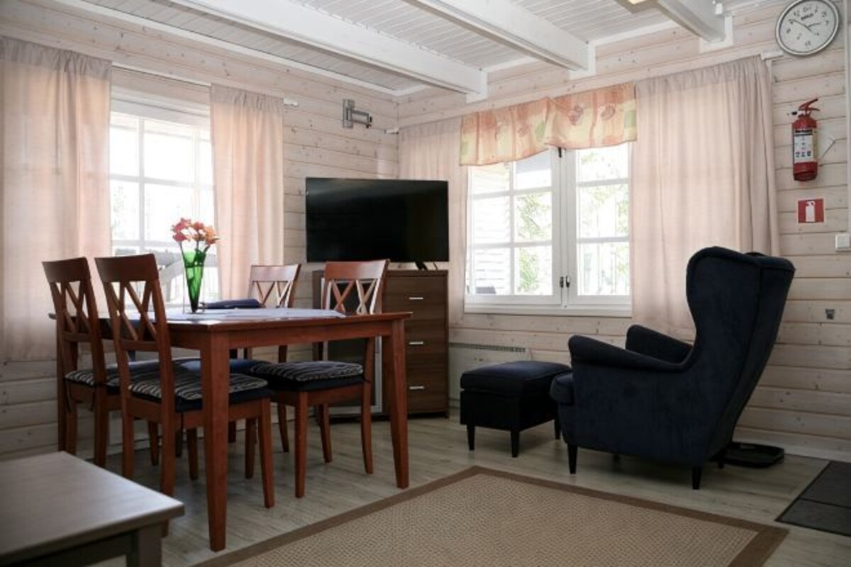 A room with a wooden dining table, chairs, a TV, a sofa, and a clock on the wall.