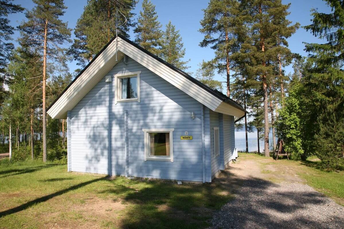 A small, light blue cabin with a gabled roof, surrounded by tall pine trees and a clear sky.