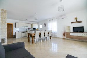 A modern living room with a sofa, dining table, chairs, a TV, and a kitchen in the background.
