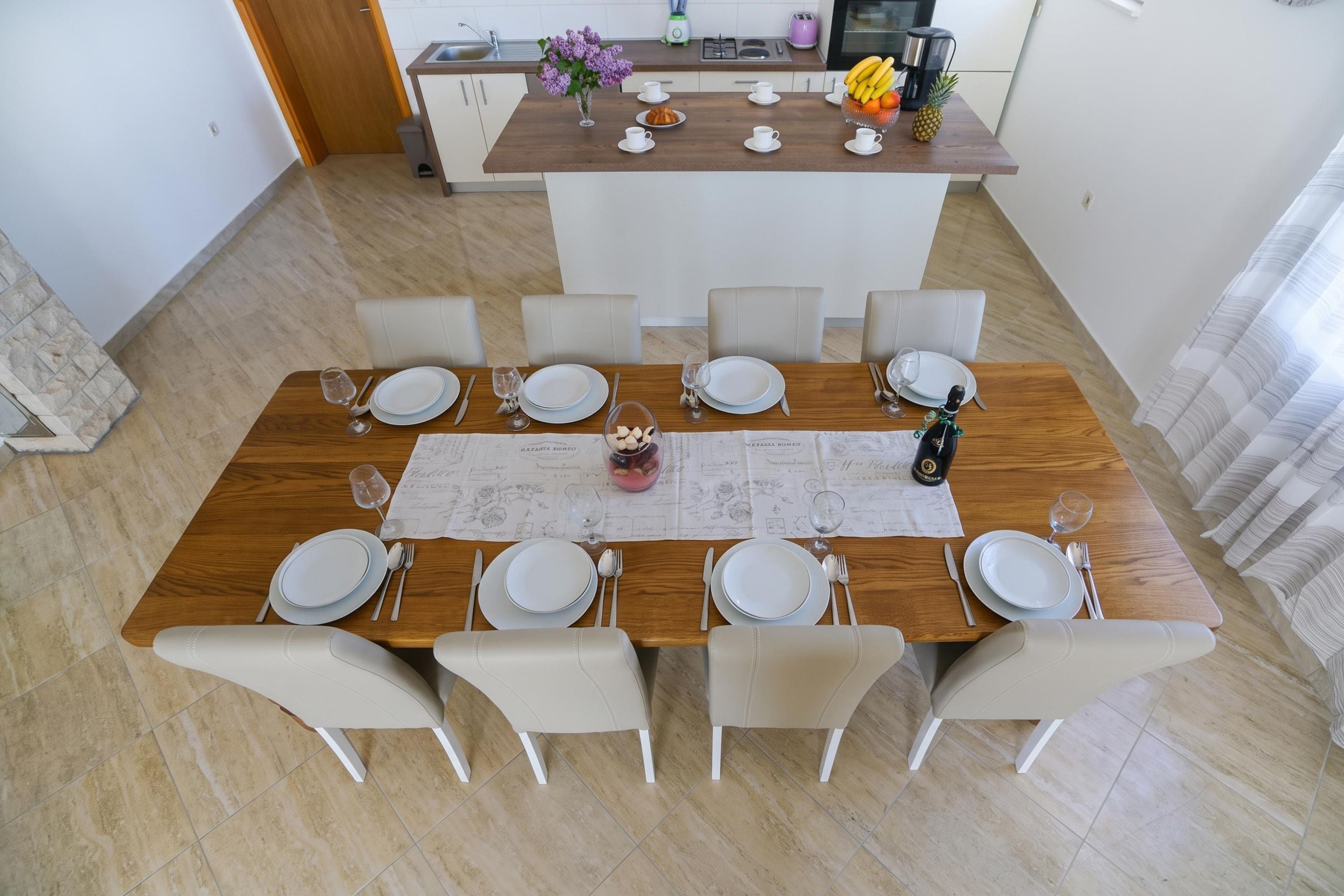 A dining area with a wooden table set for six, surrounded by white chairs, in a modern apartment with a kitchen in the background.
