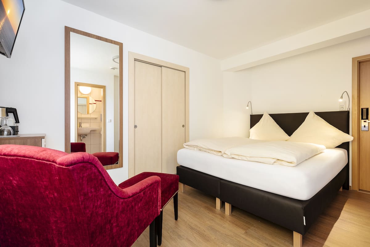 Quarto de hotel com uma cama, uma cadeira vermelha, um espelho e uma porta de madeira.