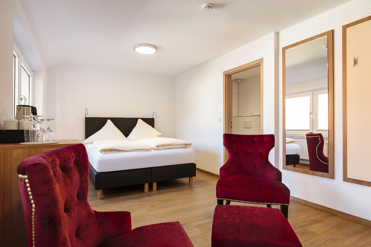 Quarto de hotel moderno com uma cama grande, uma escrivaninha de madeira, uma poltrona vermelha e um espelho grande na parede.