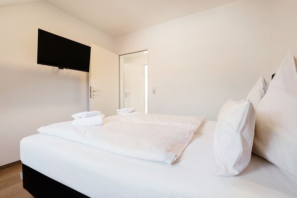 Una cama bien hecha con sábanas blancas, un televisor de pantalla plana montado en la pared y una puerta que da a otra habitación.