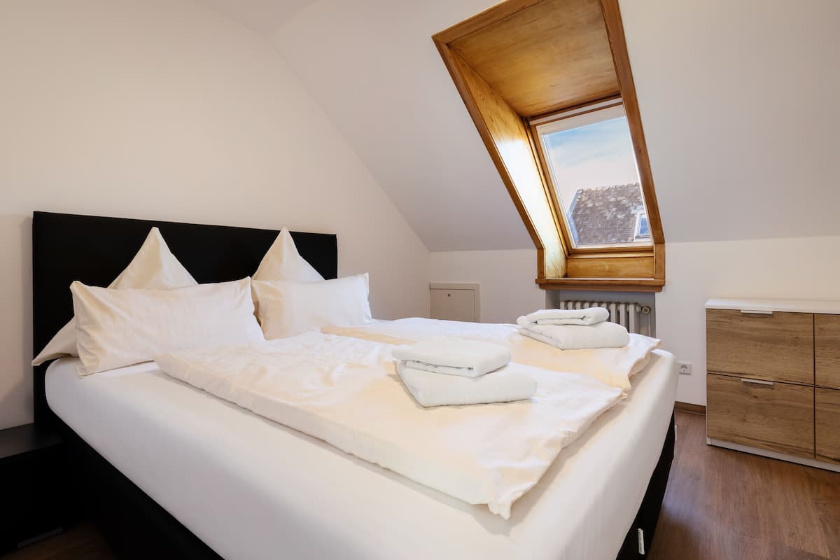 Cama bem arrumada com lençóis e almofadas brancos, cabeceira de madeira, pequena janela com vista e cômoda de madeira.