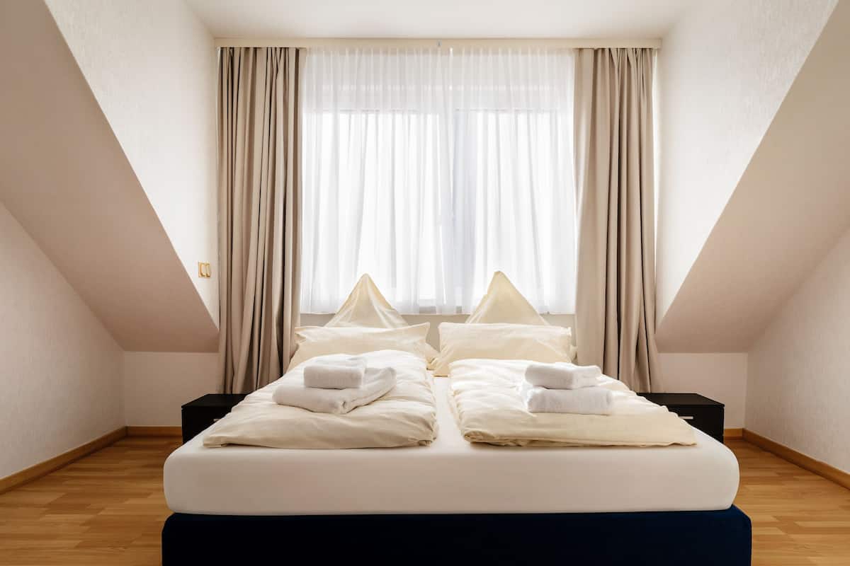 Quarto de hotel com cama, duas mesinhas de cabeceira, janela com cortinas e piso de madeira.
