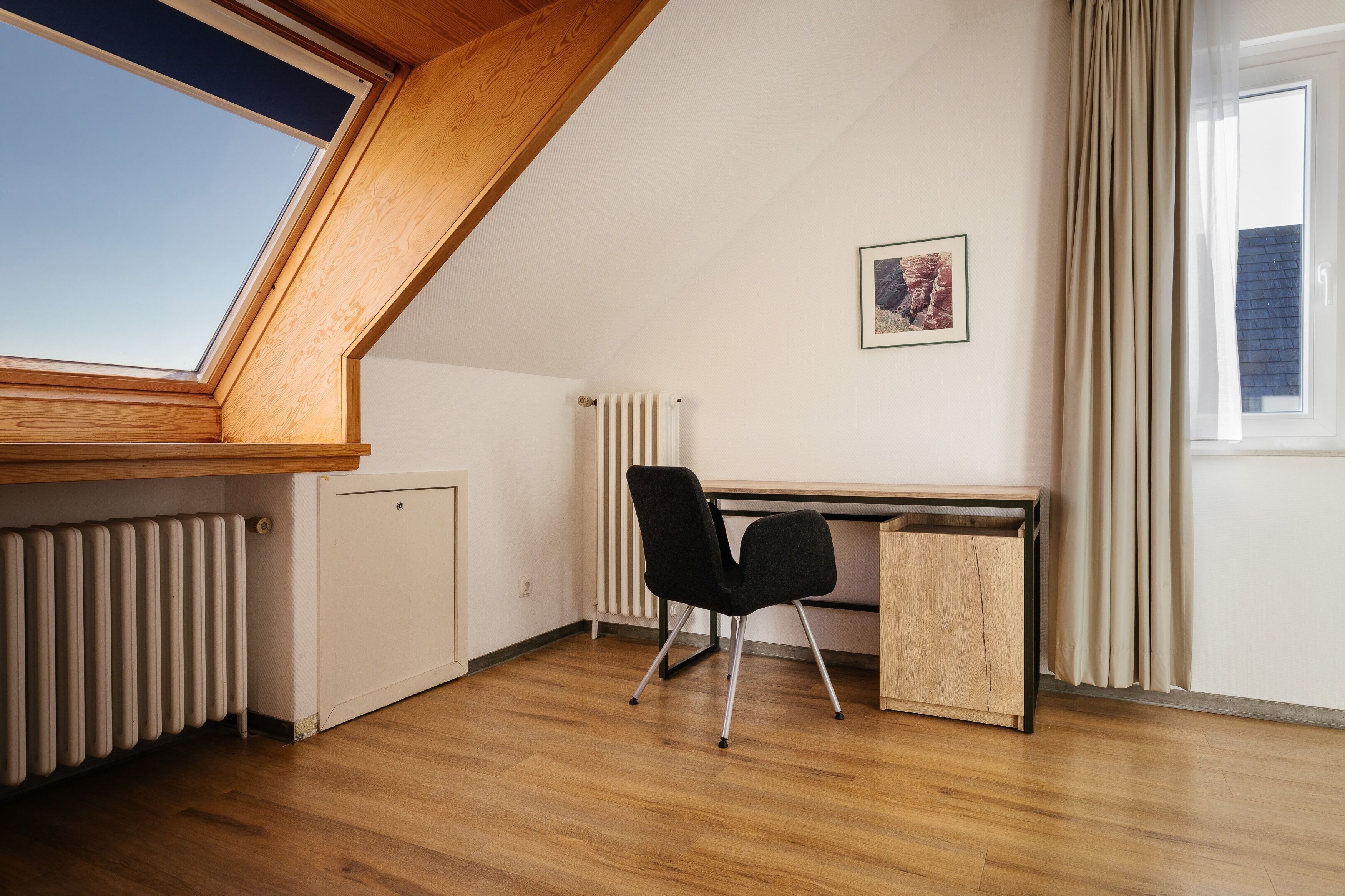 Una habitación con techo inclinado, un radiador, un escritorio con silla y una ventana con cortinas.