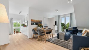 A modern living room with a dark blue sofa, a wooden dining table with chairs, and a large window with curtains. - Hohe Wacht – for 6 people, rural, idyllic, North Sea (Emmelsbüll)