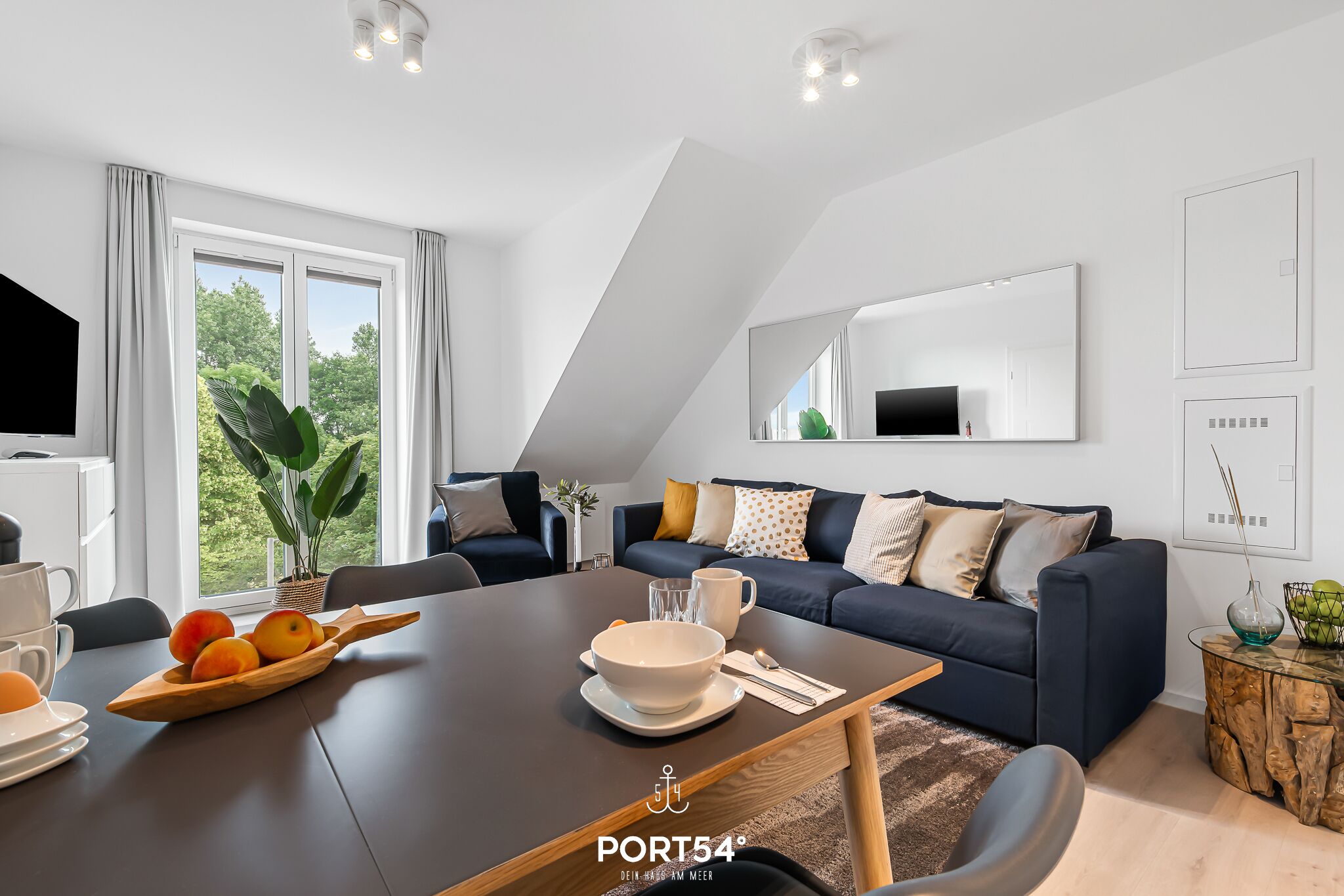 A modern living room with a dark sofa, a dining table with fruit, and a large window.