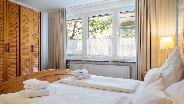 A hotel room with a wooden wardrobe, a bed with white linens, a window with curtains, and a bedside lamp. - Hummerklippen Ferienwohnungen (Helgoland)