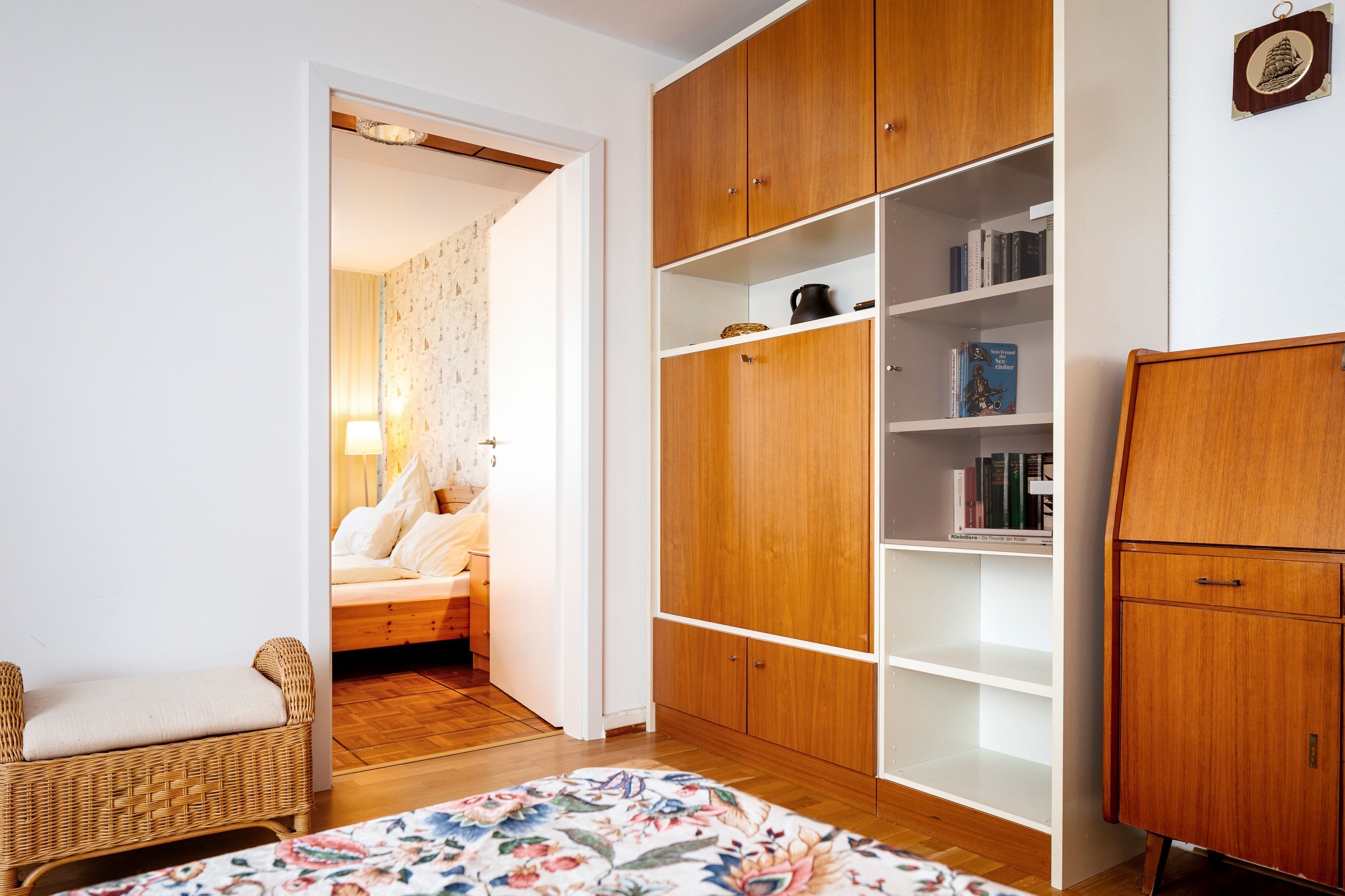 Una habitación con un armario de madera, una silla de mimbre, una cama con edredón floral y una estantería con libros.