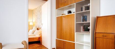 Una habitación con un armario de madera, una silla de mimbre, una cama con edredón floral y una estantería con libros.
