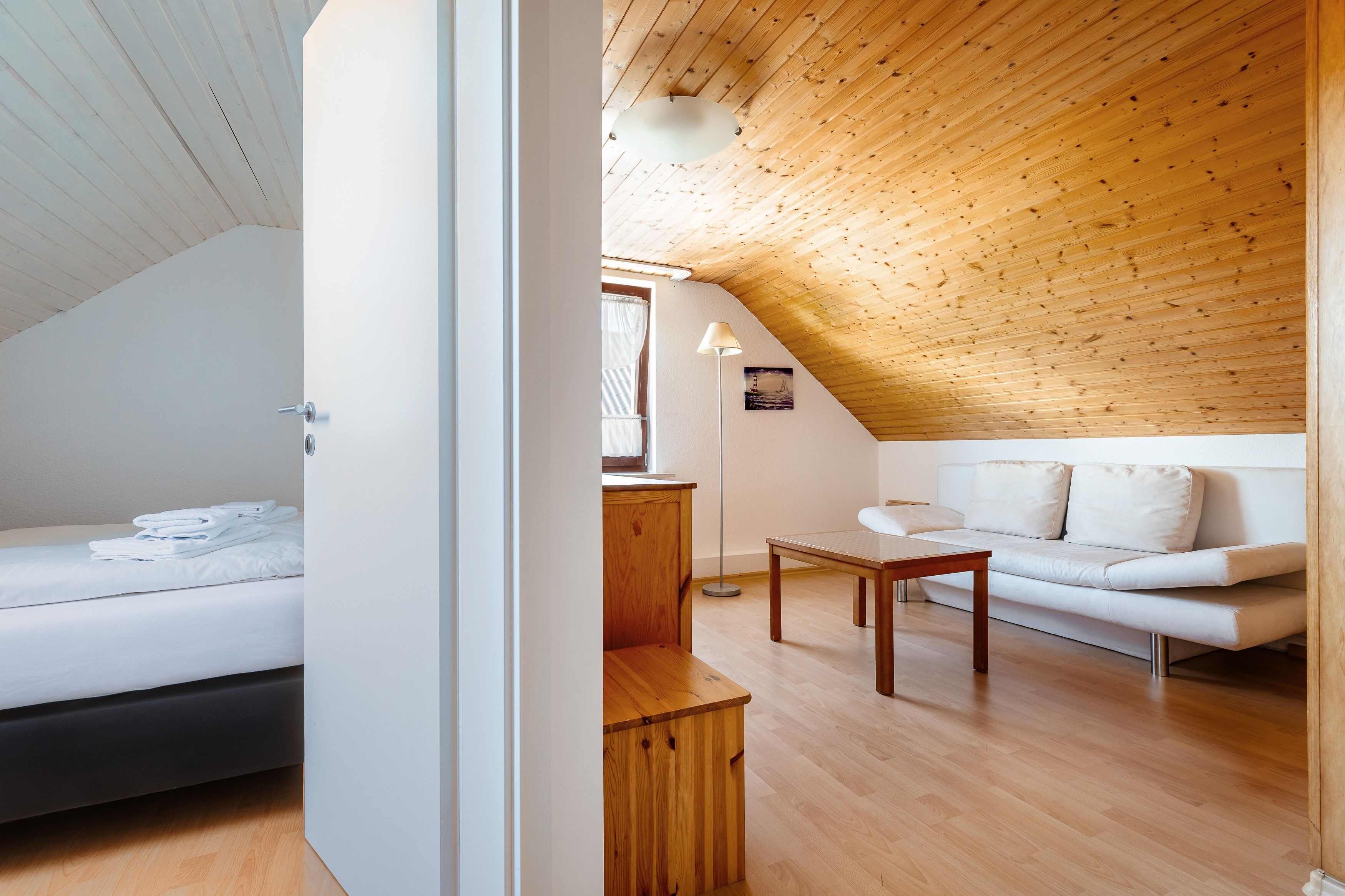 A modern, well-lit room with a wooden ceiling, a white sofa, a small wooden table, and a neatly made bed.
