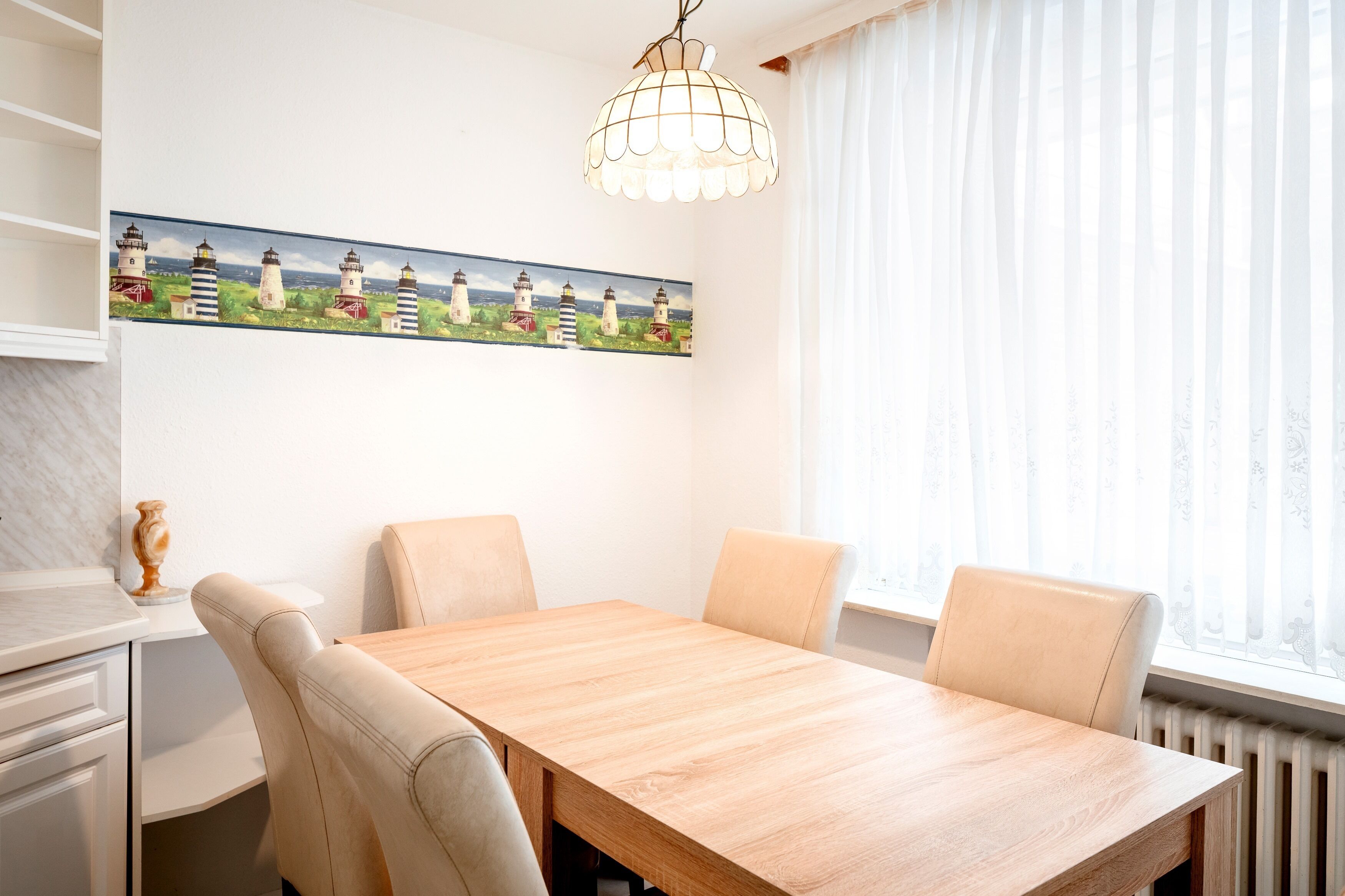 Un comedor con una mesa de madera y cuatro sillas, un cuadro de faros en la pared, un armario blanco y una ventana con cortinas translúcidas.