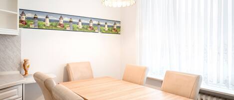 Un comedor con una mesa de madera y cuatro sillas, un cuadro de faros en la pared, un armario blanco y una ventana con cortinas translúcidas.