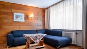 A modern living room with a blue sofa, a small wooden table with a floral tablecloth, a picture on the wall, and a window with sheer curtains. - Hummerklippen Ferienwohnungen (Helgoland)