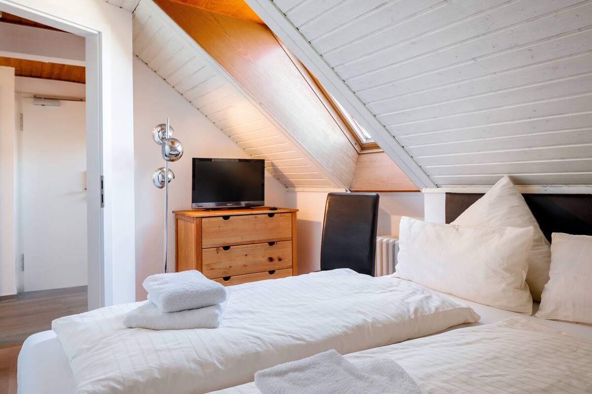 Ein Schlafzimmer mit schrägen Wänden, einer Holzkommode, einem Fernseher und einem Bett mit weißer Bettwäsche und Kissen.