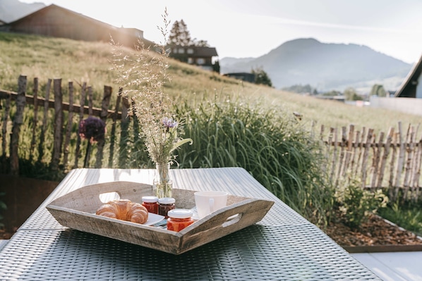 Un plateau en bois avec des croissants et de la confiture, posé sur une table en métal à l’extérieur, entouré d’une clôture en bois et de verdure, avec des montagnes en arrière-plan.