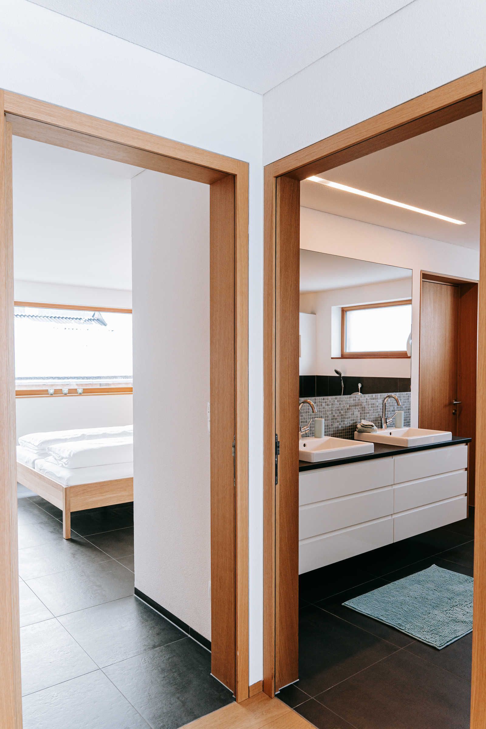A modern bathroom with a double sink, a mirror above the sink, and a wooden door leading to a bedroom with a bed and a window.