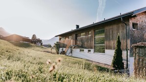 A modern house with a wooden exterior, a staircase leading to a deck, and a view of the surrounding landscape. - Baldauf Apartment - Apartment (Hittisau)