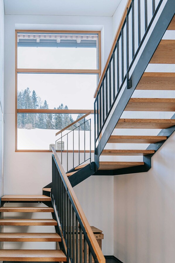 A modern staircase with wooden steps and metal railings, leading to a room with a large window offering a view of a snowy landscape.