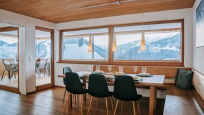A dining area with a wooden table, four chairs, and large windows offering a mountain view. - Baldauf Apartment - Apartment (Hittisau)