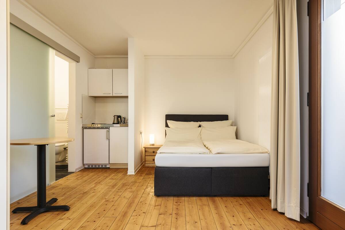 Un dormitorio con cama, una mesita de noche, un escritorio pequeño y una kitchenette al fondo.