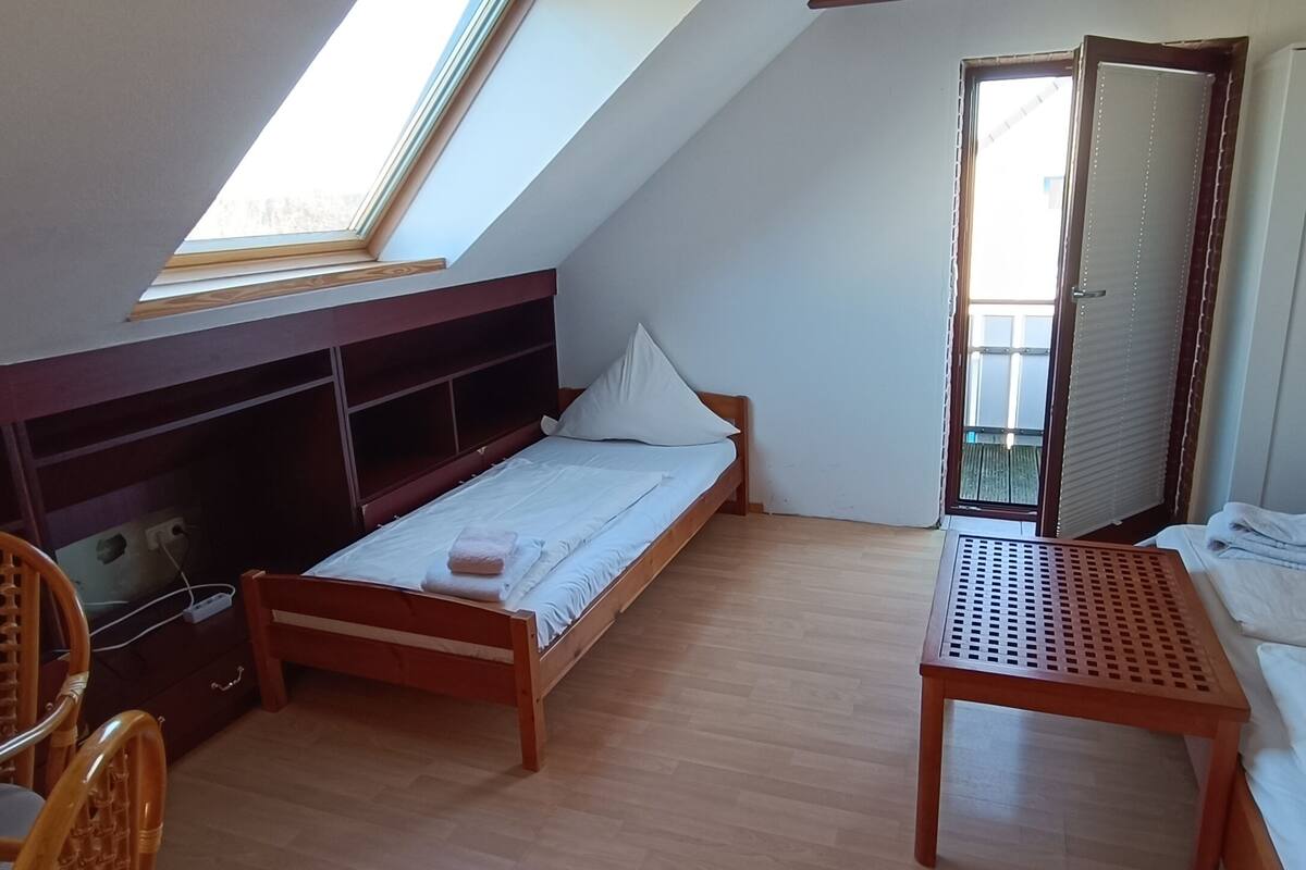 Una habitación pequeña y bien iluminada, con techo inclinado, una cama individual, un escritorio de madera, una silla y un armario.