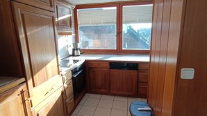 A compact kitchen with wooden cabinets, a dishwasher, and a window overlooking a residential area. - Haus Hoog Stean (Helgoland)
