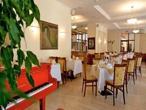 Une salle à manger avec des tables dressées pour le service, un piano et un grand tableau accroché au mur.