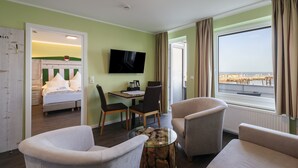 A hotel room with a bed, a TV, a dining table with chairs, a coffee table, and a large window with a view of the sea. - Insulaner Ferienwohnungen (Helgoland)