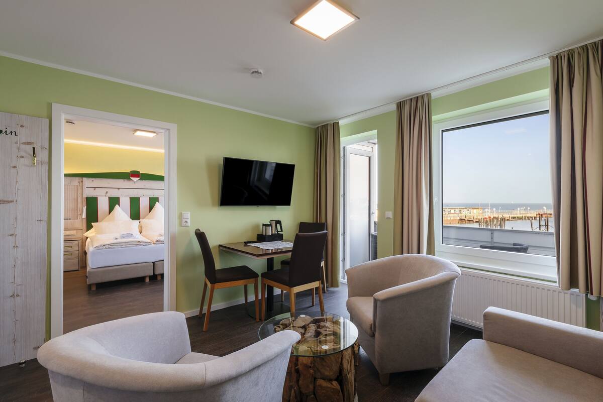 A hotel room with a bed, a TV, a dining table with chairs, a coffee table, and a large window with a view of the sea.