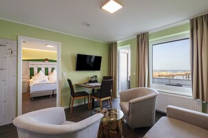 Habitación de hotel con cama, televisión, mesa de comedor con sillas, mesa de centro y un amplio ventanal con vistas al mar.