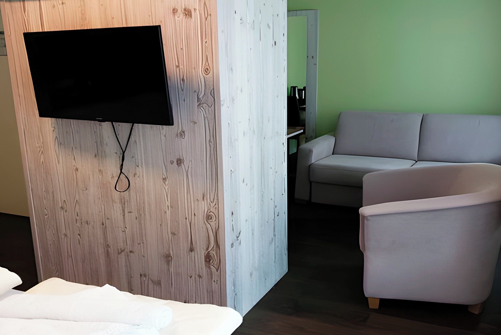 A room with a flat-screen TV mounted on a wooden wall, a grey sofa, and a white armchair.