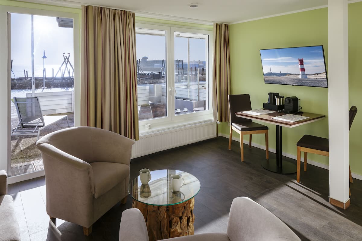 A hotel room with a large window, a glass table, two chairs, a coffee maker, and a television on the wall.