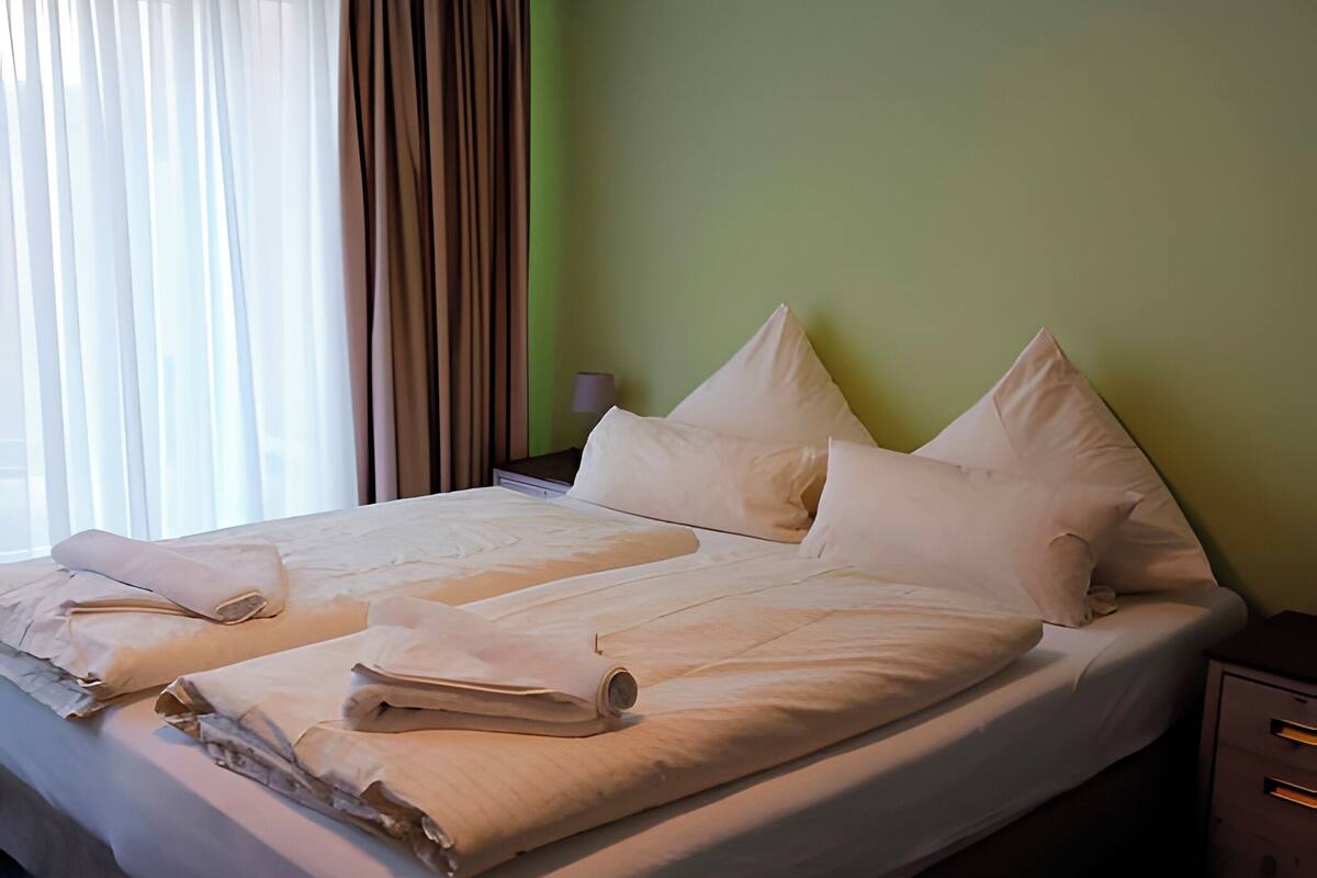 A neatly made bed with white linens and pillows, a bedside lamp, and a nightstand with drawers.