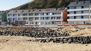 A row of modern, multi-story buildings with a flat roof and solar panels, situated on a sandy beach with a rocky foreground. - Insulaner Ferienwohnungen (Helgoland)