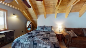 Quarto com teto inclinado, cama com colcha estampada, mesa de cabeceira, luminária, espelho, cômoda e sofá de couro.