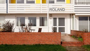 A two-story building with a yellow and white facade, a nameplate reading "ROLAND," and a brick wall in the foreground. - Haus Roland (Helgoland)