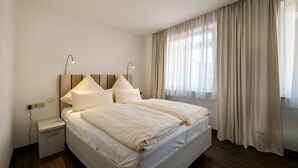 A neatly made bed with white linens, a bedside table with a lamp, and a window with curtains. - Haus Roland (Helgoland)