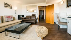 A modern living room with a sofa, a coffee table, and a unique woven ottoman. There is a dining area with a chair and a table, and a wooden door leading to another room. - Haus Roland (Helgoland)