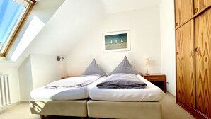 A bedroom with a skylight, a bed with striped bedding, a nightstand with a lamp, and a wooden wardrobe. - Haus Germania (Helgoland)