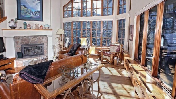 A spacious living room with a fireplace, leather sofa, wooden coffee table, and large windows offering a view of trees. - Beaver Creek Pines 4 Bedroom Ski in, Ski out Vacation Rental With Restaurant, Outdoor Pool, Jacuzzi and Fitness Center (Avon)