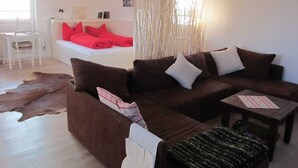 Een woonkamer met een bruine bank, een klein tafeltje, een witte stoel en een bed met rode kussens.