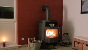 Een kamer met een houtkachel, een mand houtblokken en een kaars op een raamdorpel.