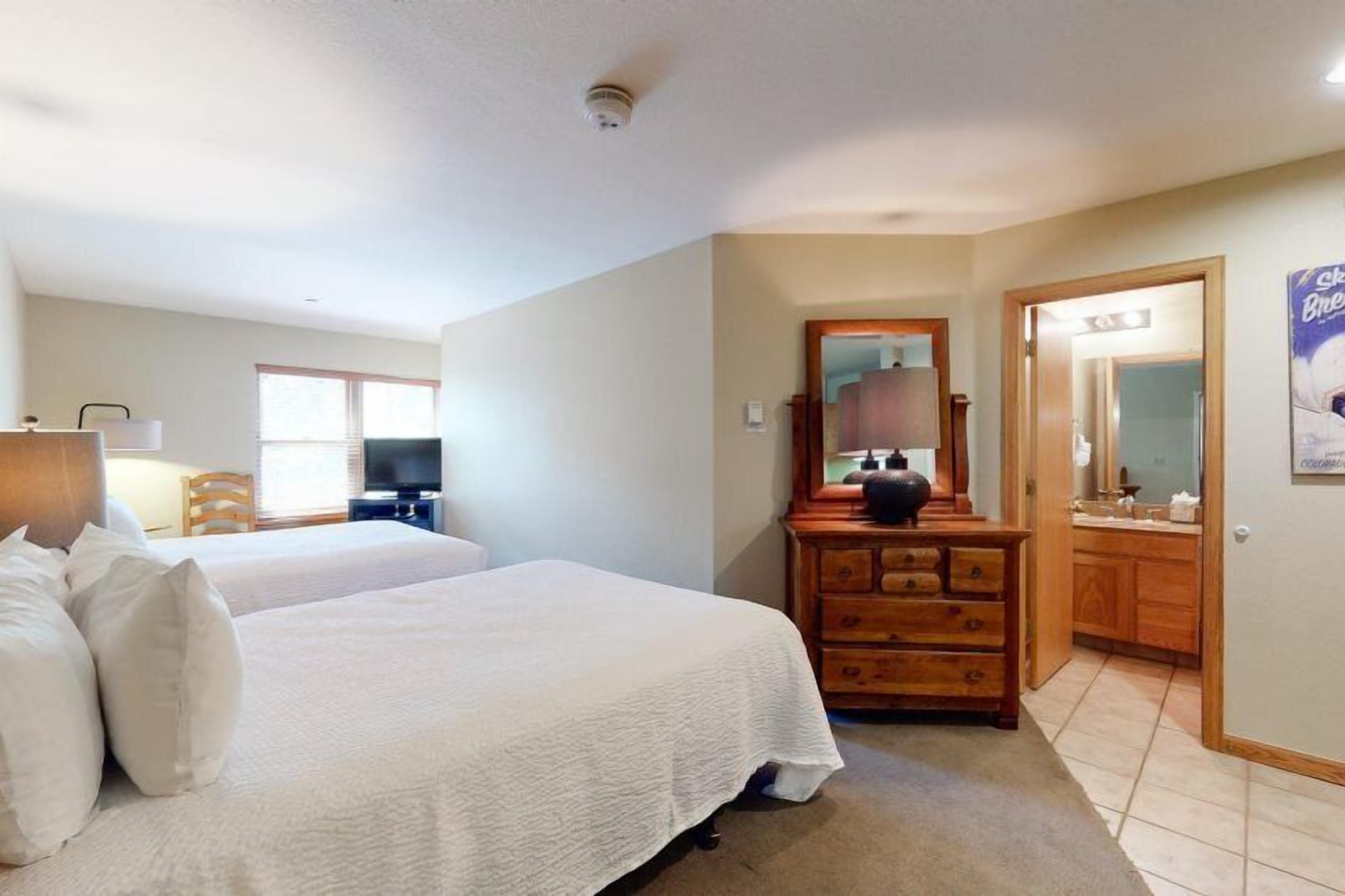 Ein Hotelzimmer mit zwei Betten, einem Holzkapfen, einem Spiegel und einem durch eine offene Tür einsehbaren Badezimmer.
