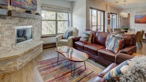 A spacious living room with a stone fireplace, leather sofas, a glass coffee table, and large windows offering natural light. - 4 Bedroom Ski in Condo Only One Block From Historic Downtown, Shopping, Restaurants and Entertainment (Breckenridge)