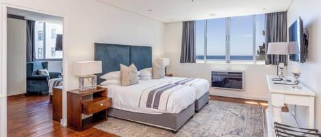 A modern hotel room with a large bed, a desk with a flat-screen TV, a seating area with a chair, and a view of the sea through the windows.