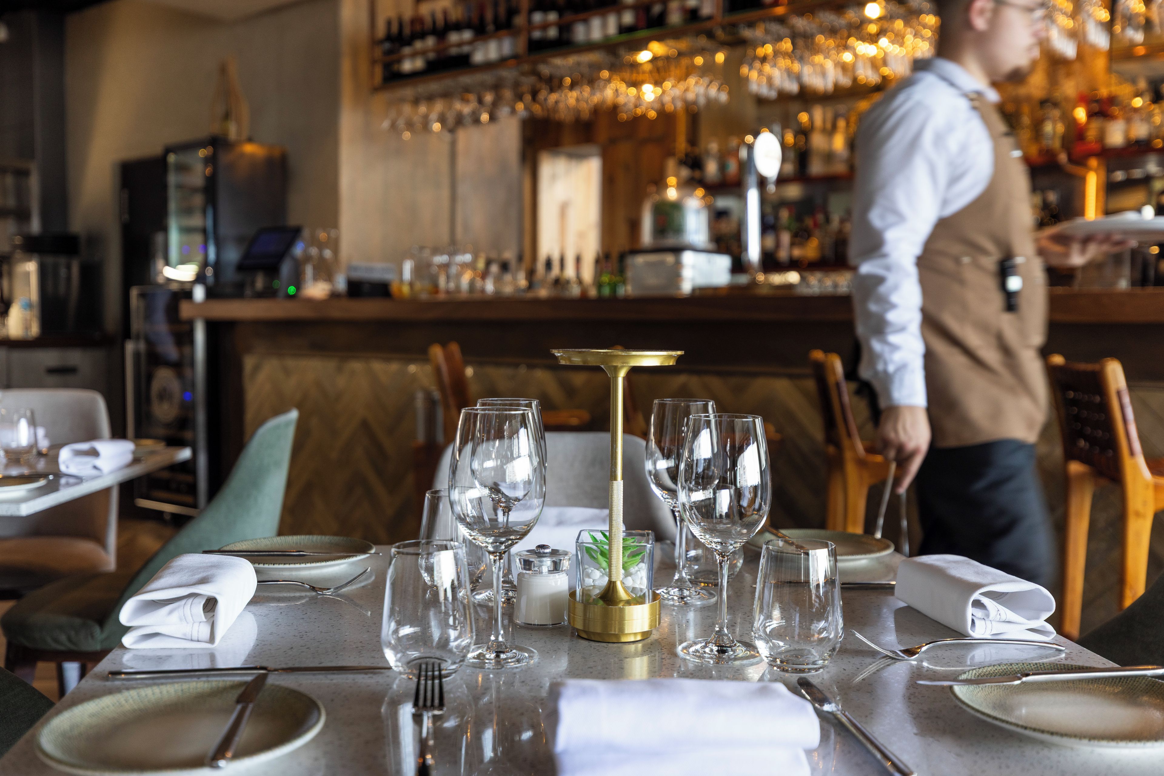 A restaurant setting with tables set for dining, a bar area in the background, and a waiter attending to a table.